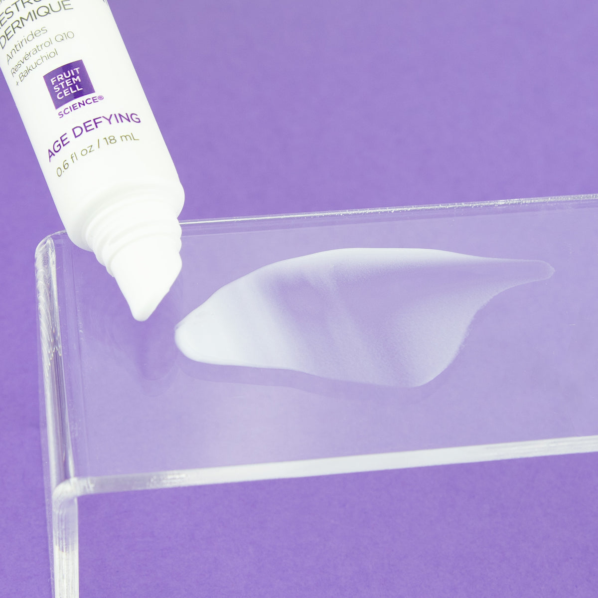 A close-up of a white, age-defying cream being dispensed from a tube onto a glass against a purple background.