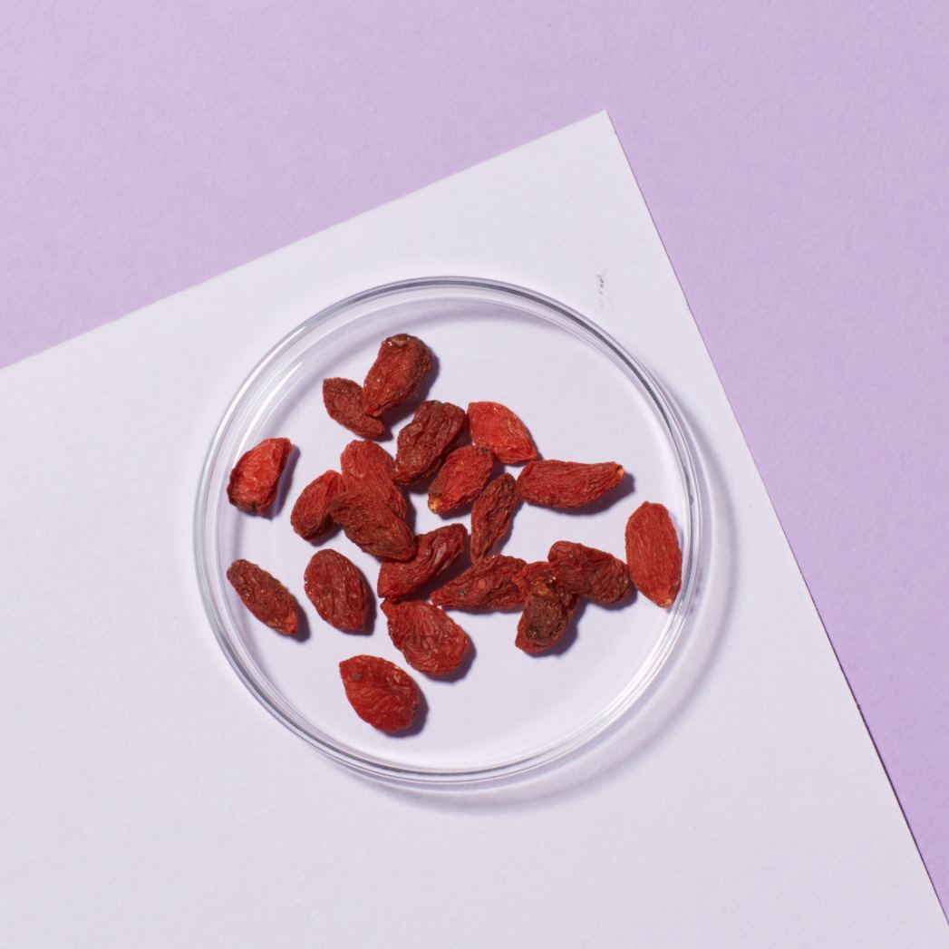 Dried goji berries placed in a clear glass dish on top of a white and lavender background.