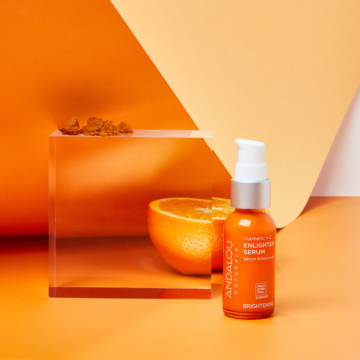 Brightening Turmeric + Vitamin C Enlighten Serum from Andalou Naturals with turmeric powder, an orange, and a glass cube on an orange background.